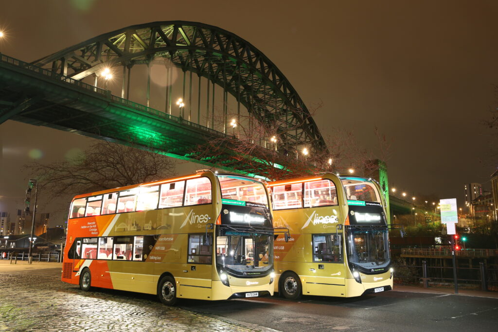 Two X Lines double decker buses sit under the Tyne Bridge at night time.